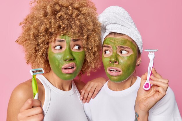 Verlegen halten zwei junge Frauen Rasierer, die Hygienemaßnahmen durchführen werden, rasieren die Beine und tragen eine grüne Schönheitsmaske aus Gurken auf, um die Haut feuchtigkeitsspendend zu tragen und ein Badetuch auf dem Kopf zu tragen, das über rosafarbenem Hintergrund isoliert ist