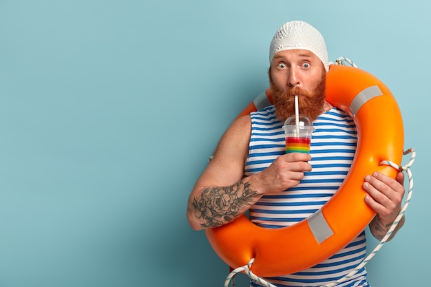 Verlegener Urlauber trinkt kalten Sommercocktail, verbringt Freizeit am Strand, trägt Swimcap Sailor T-Shirt