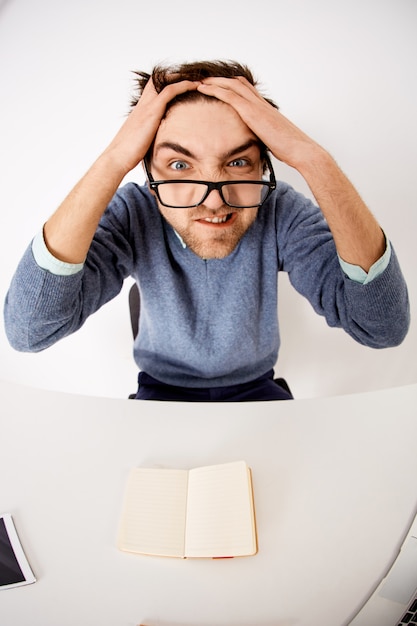 Verrückter und verzweifelter junger unter Druck stehender Mann, zerzauste Haare und grimmig verzogenes Gesicht bei der Arbeit, sitzen Schreibtisch kann sich keine Ideen ausdenken