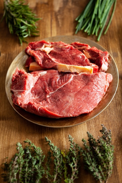 Verschiedene Arten von rotem Fleisch in einem Vintage-Teller neben grünem Gemüse und Kräutern auf Holztisch. Zubereitung des Abendessens.