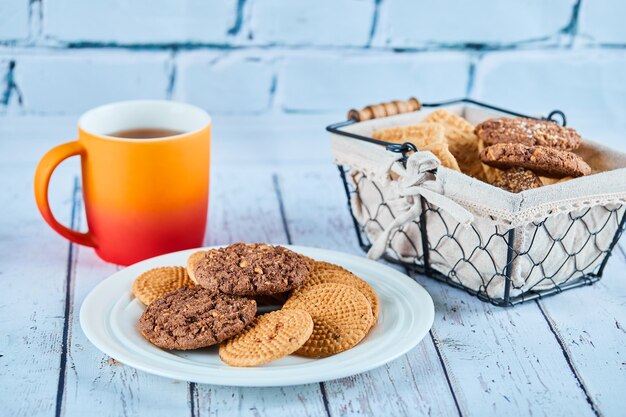 Verschiedene Kekse in Teller und Korb und eine Tasse auf blauem Tisch.