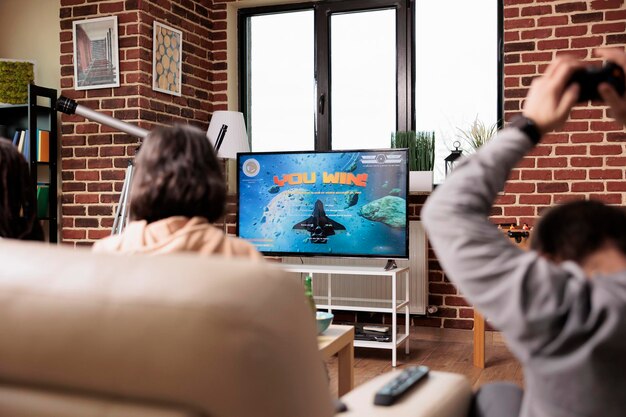 Verschiedene Leute sitzen im Wohnzimmer und genießen Spiele auf elektronischen Unterhaltungsgeräten. Verspielte multiethnische Gruppe von Freunden, die auf dem Sofa sitzen, während sie Konsolen-Videospiele mit Controller spielen.