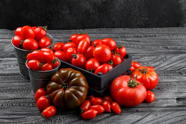 Verschiedene Tomaten in Holzkiste, Mini-Eimer auf grauer Holz- und dunkler Wand, hohe Winkelansicht.