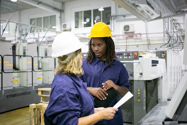 Verschiedene weibliche Fabrikangestellte in Schutzhelmen und Overalls, die auf dem Werksgelände stehen und sich unterhalten