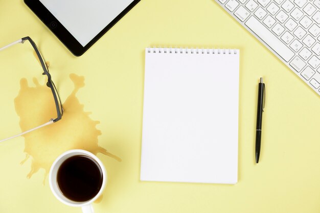Verschütteter Kaffee; Brille; digitales Tablett; Tastatur; leerer gewundener Notizblock und Stift auf gelbem Hintergrund