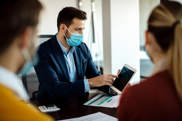 Versicherungsagent mit Gesichtsmaske mit Touchpad bei einem Treffen mit Kunden im Büro