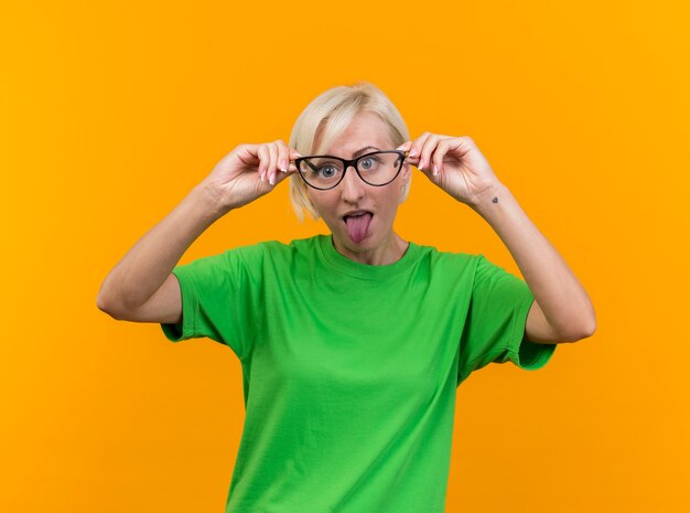 Verspielte blonde slawische Frau mittleren Alters, die Kamera betrachtet, die Brille vor Augen hält, die Zunge lokalisiert auf gelbem Hintergrund zeigt