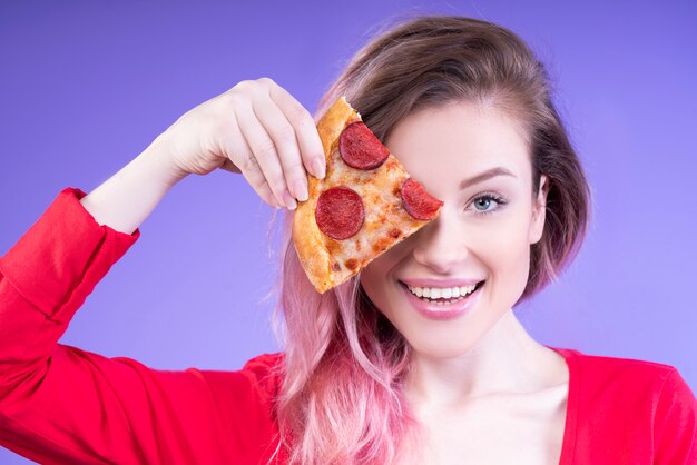 Verspielte Frau, die ein Stück Pizza nahe ihrem rechten Auge hält