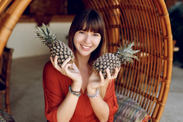 Verspielte Frau, die zwei Ananas hält und auf hängendem Bambusstuhl sitzt