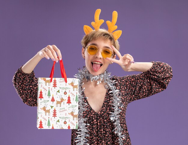 Verspielte junge hübsche Frau, die Rentiergeweih-Stirnband und Lametta-Girlande um den Hals mit Gläsern hält, die Weihnachtsgeschenktüte halten