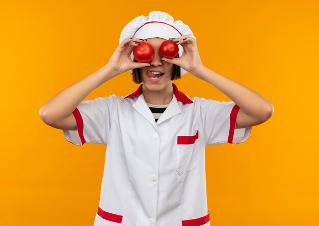 Verspielte junge Köchin in der Kochuniform, die Tomaten auf Augen setzt und Zunge lokalisiert auf orange Hintergrund zeigt