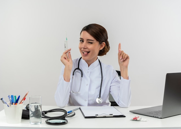Verspielte junge Ärztin, die medizinische Robe und Stethoskop trägt, sitzt am Schreibtisch mit medizinischen Werkzeugen und Laptop, die Spritze halten, die Zunge zeigt und Finger lokalisiert hebt