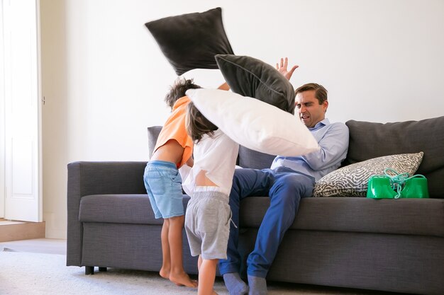 Verspielte Jungen, die kaukasischen Mann mit Kissen angreifen. Fröhliche Kinder spielen und haben Spaß mit seinem Vater. Papa schließt die Augen und verteidigt sich vor Angriffen. Konzept für Kinder-, Familien- und Spielaktivitäten