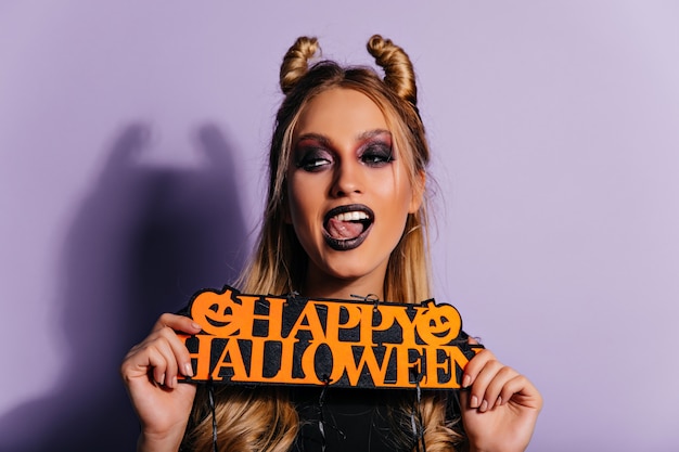 Verspielte kaukasische junge Frau, die Halloween-Fotoshooting genießt. Blondes Mädchen in der Vampirkleidung, die mit Partydekor aufwirft.