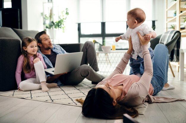 Verspielte Mutter, die ihren kleinen Sohn hält, während sie im Wohnzimmer auf dem Boden liegt Vater und Tochter benutzen Laptop