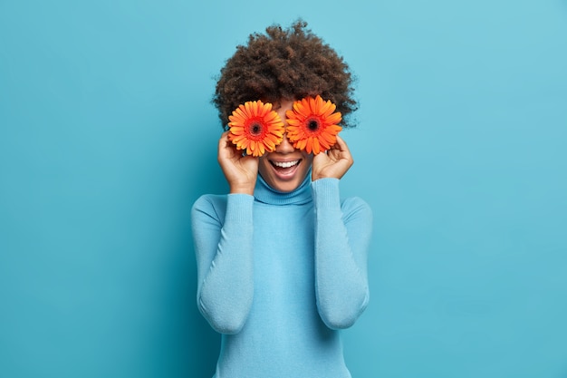 Verspielte positive Afroamerikanerin bedeckt die Augen mit zwei orangefarbenen Gerbera, genießt den Frühling, frische Blumen, hat Spaß, ist lässig gekleidet,