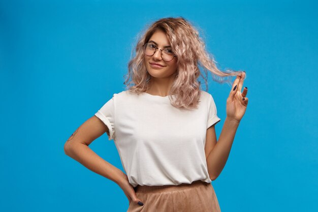 Verspielte Studentin in übergroßem weißem T-Shirt und runder Brille, mit flirtendem Lächeln, das ihr rosa Haar um den Finger dreht. Menschen, Lebensstil, Weiblichkeit