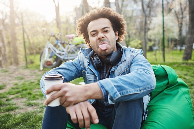 Verspielter attraktiver Afroamerikaner mit Afro-Frisur, die Zunge herausstreckt und kindisch ist, während auf schlechtem Bohnenstuhl mit Tasse Kaffee sitzend