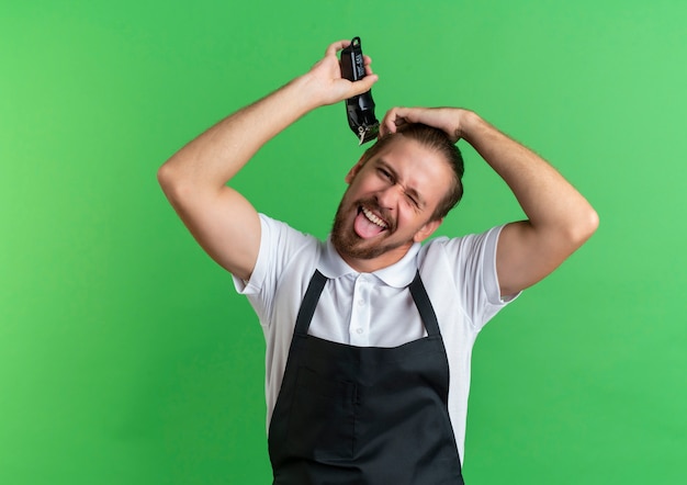 Kostenloses Foto verspielter junger hübscher friseur, der uniform trägt, die sein haar mit hand auf kopf zwinkert und zunge isoliert auf grünem hintergrund mit kopienraum zeigt