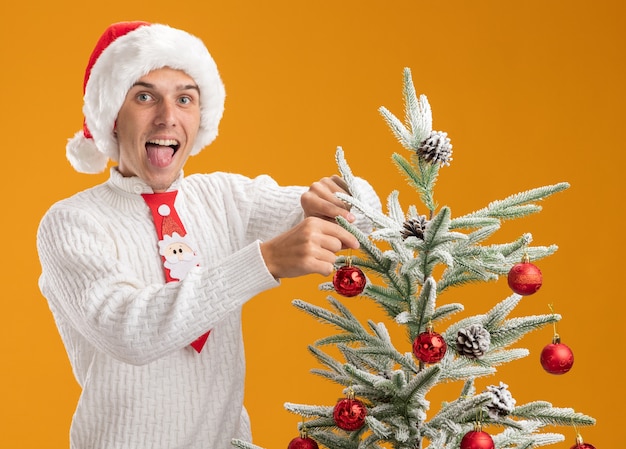 Verspielter junger hübscher Kerl, der Weihnachtshut und Weihnachtskrawatte trägt, die nahe Weihnachtsbaum steht, der sie mit Weihnachtsballverzierung verziert