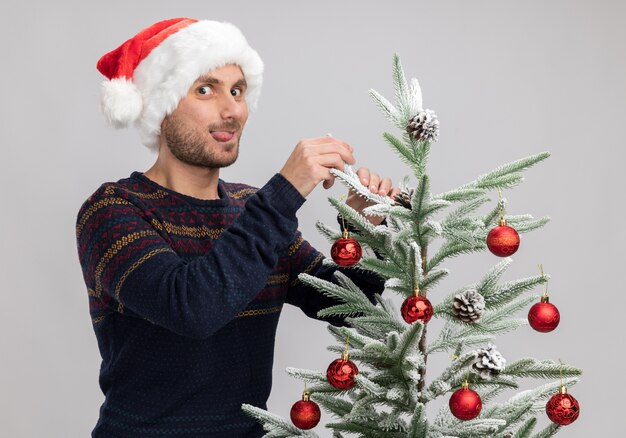 Verspielter junger kaukasischer Mann, der Weihnachtshut trägt, der nahe Weihnachtsbaum berührt, der Kamera betrachtet, die Zunge lokalisiert auf weißem Hintergrund zeigt