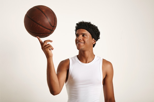 verspielter lächelnder junger afroamerikanischer Basketballspieler, der ein weißes ärmelloses Hemd trägt, das einen alten Lederball auf einem Finger lokalisiert auf Weiß hält.