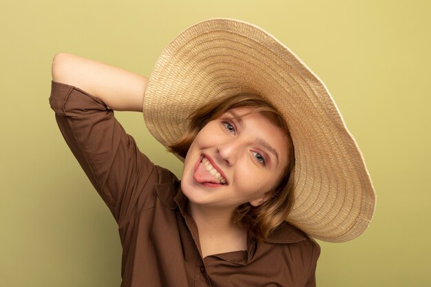 Verspieltes junges blondes Mädchen mit Strandhut, das den Kopf berührt und Zunge zeigt