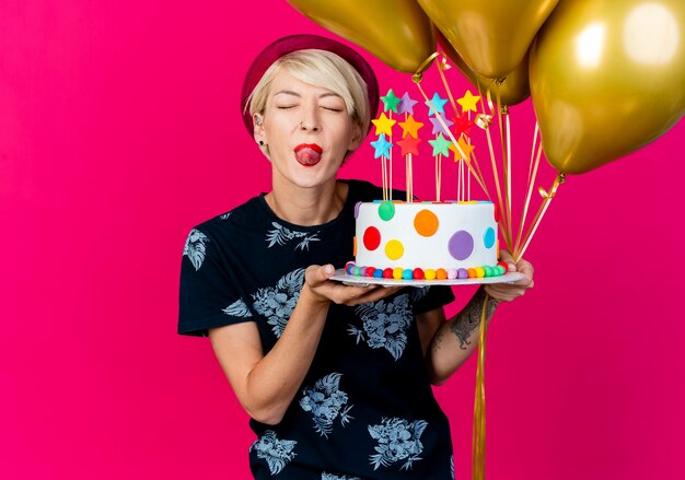 Verspieltes junges blondes Partygirl, das Partyhut hält, der Luftballons und Geburtstagstorte mit Sternen hält, die Zunge mit geschlossenen Augen lokalisiert auf purpurrotem Hintergrund mit Kopienraum zeigen