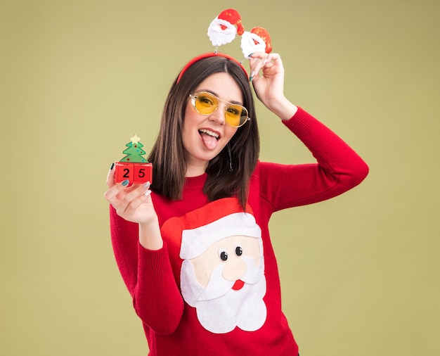 Verspieltes junges hübsches kaukasisches Mädchen, das Weihnachtsmann-Pullover und Stirnband mit Brille trägt, die Weihnachtsbaumspielzeug mit Datum halten