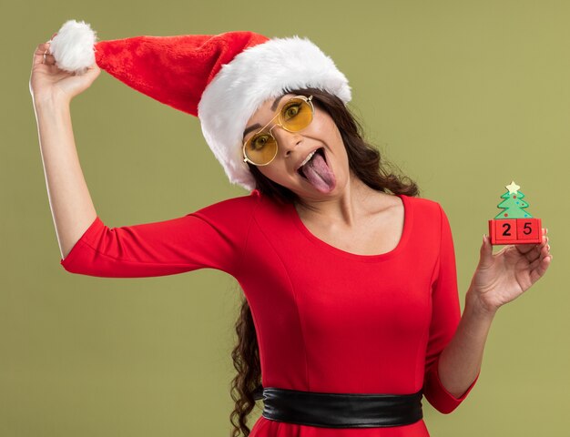 Verspieltes junges hübsches Mädchen mit Weihnachtsmütze und Brille, das Weihnachtsbaumspielzeug mit Date-Grabing-Hut hält, der Zunge isoliert auf olivgrüner Wand zeigt