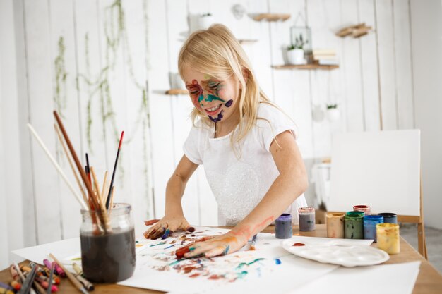 Verspieltes, süßes blondes Mädchen, das Spaß hat, indem es Bild mit ihren Händen zeichnet, ihre Handflächen in verschiedenen Farben vertieft und sie auf weißes Blatt Papier legt.