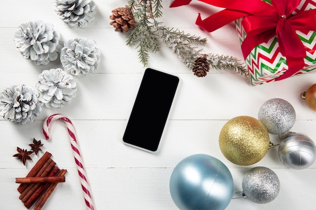 Verspotten Sie den leeren leeren Bildschirm des Smartphones an der weißen Holzwand mit farbenfroher Feiertagsdekoration und Geschenken.