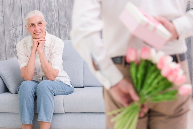 Versteckender Blumenstrauß und Geschenkbox des Defocused Mannes vor seiner lächelnden Frau