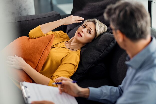 Verstörte Frau, die sich auf die Couch des Psychiaters legt und mit ihrem Therapeuten spricht