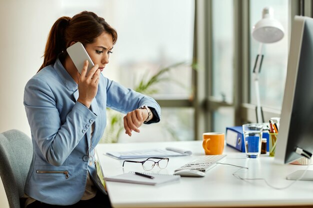 Verstörte Geschäftsfrau, die am Telefon spricht, während sie auf ihrer Armbanduhr im Büro die Zeit ansieht