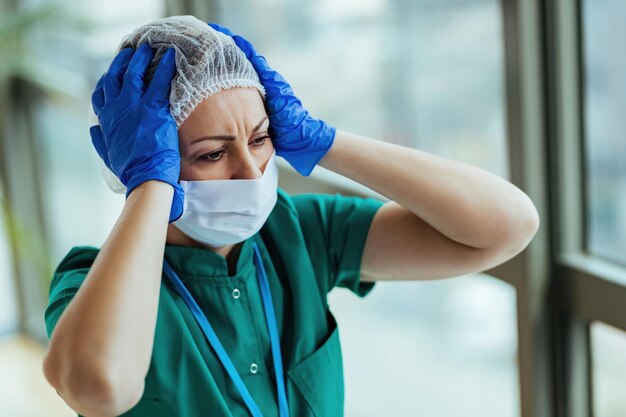 Verstörte Krankenschwester, die Schutzkleidung trägt, während sie ihren Kopf in der medizinischen Klinik vor Schmerzen hält