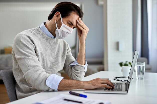 Verstörter Geschäftsmann mit schützender Gesichtsmaske, der seinen Kopf vor Schmerzen hält, während er zu Hause an einem Computer arbeitet