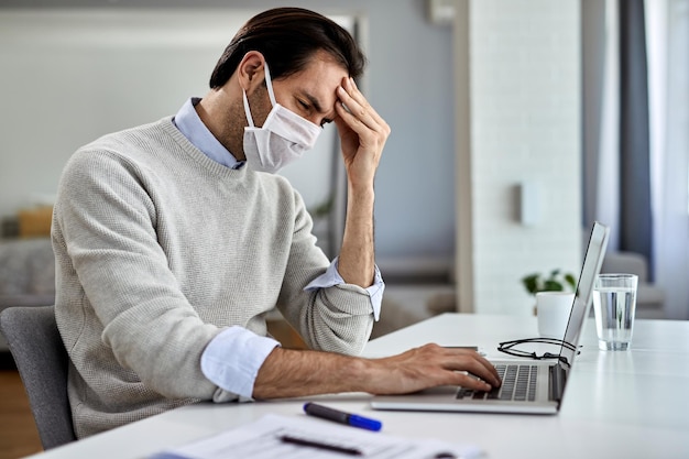 Verstörter Geschäftsmann mit schützender Gesichtsmaske, der seinen Kopf vor Schmerzen hält, während er zu Hause an einem Computer arbeitet