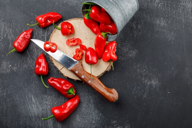 Verstreute rote Paprika mit Holzstück, Messer in einem Mini-Eimer an grungy grauer Wand, flach liegen.