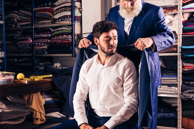 Versuchender Mantel des älteren männlichen Modedesigners über seinem Kunden im Shop