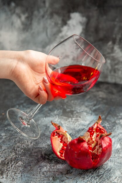 Vertikale Ansicht der Frauenhand, die ein Glas trockenen Rotweins und einen offenen Granatapfel auf grauem Hintergrund hält