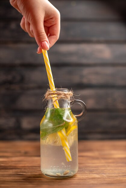 Vertikale ansicht der hand, die ein rohr in einem glas mit frischem detox-wasser auf braunem hintergrund hält