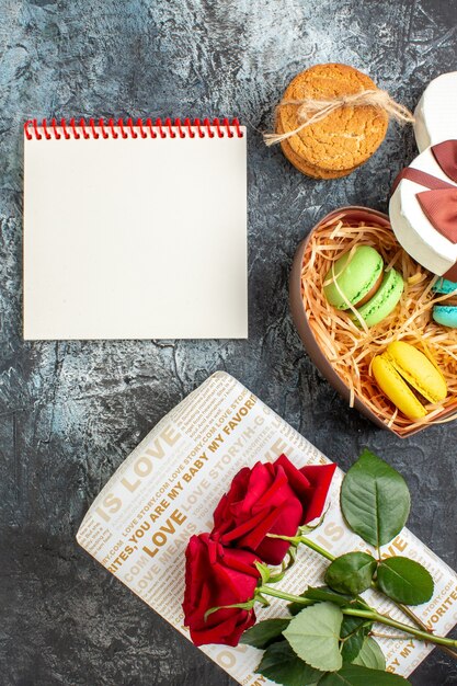 Vertikale Ansicht der herzförmigen schönen Geschenkbox mit köstlichen Macarons und Keksen, rote Rose Spiralnotizbuch auf eisigem dunklem Hintergrund