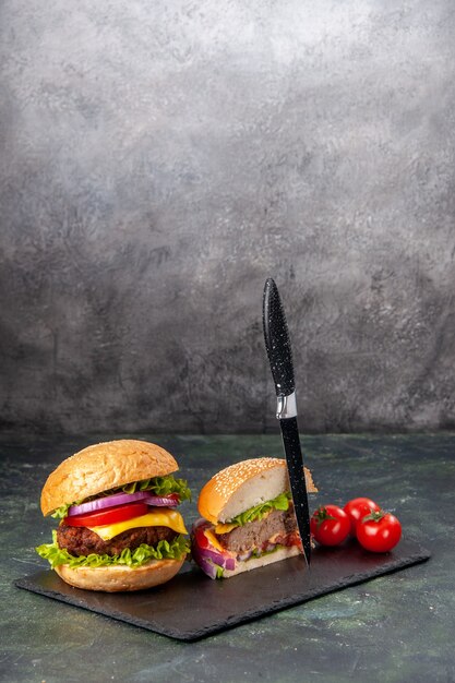 Vertikale Ansicht des ganzen Schnitts verschiedene leckere Sandwiches und Tomaten mit Stielmesser auf schwarzem Tablett auf dunkler Mischfarboberfläche