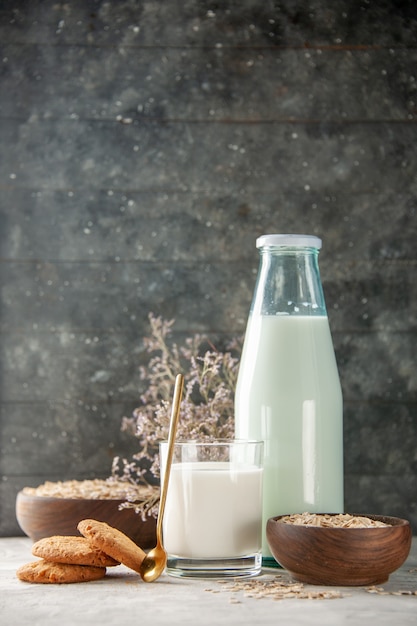 Vertikale Ansicht des Glasflaschenbechers gefüllt mit Milch und goldenen Löffelplätzchen Blumenhafer innerhalb und außerhalb des braunen Topfes auf grauem Tisch auf dunklem Holzhintergrund