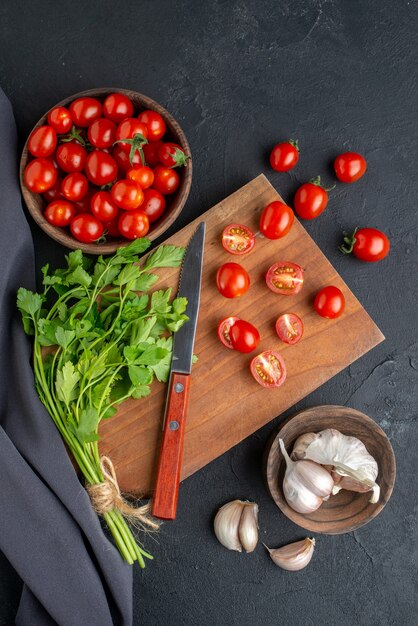 Vertikale Ansicht des grünen Bündels frischer Tomaten auf hölzernem Schneidebrett und im Schüssel Knoblauchhandtuch auf schwarzer notleidender Oberfläche