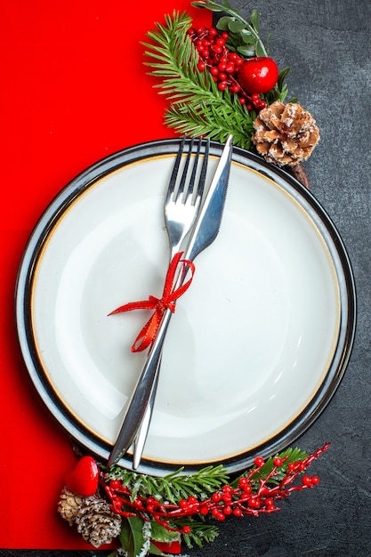 Kostenloses Foto vertikale ansicht des weihnachtshintergrundes mit besteck, das mit rotem band auf einem teller-dekorationszubehör eingestellt wird, tannenzweige auf einer roten serviette