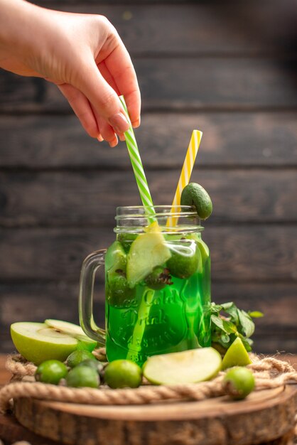 Vertikale Ansicht von frischem, köstlichem Fruchtsaft, serviert mit Apfel- und Feijoas-Hand, die Rohr auf einem Holzbrett auf einem braunen Tisch hält