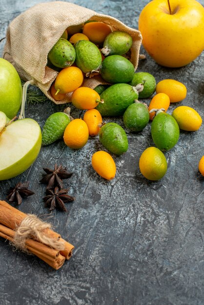 Vertikale Ansicht von frischen Kumquats innerhalb und außerhalb einer gefallenen kleinen weißen Tasche neben Zitronen- und Apfelscheiben Zimtlimetten auf grauem Hintergrund