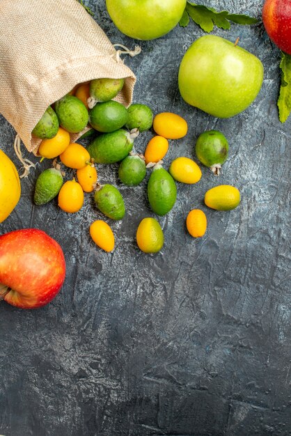 Vertikale Ansicht von frischen Kumquats innerhalb und außerhalb einer gefallenen kleinen weißen Tasche und rot-gelb-grünen Äpfeln auf grauem Tisch